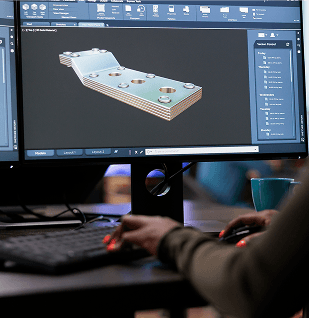 Esta imagem mostra uma pessoa trabalhando em uma estação de computador com dois monitores. No monitor principal, em primeiro plano, exibe-se um software de design 3D com uma representação detalhada de um componente metálico complexo, possivelmente uma placa com múltiplos orifícios e canais. O software mostra diversas ferramentas e menus típicos de CAD (Computer-Aided Design). A pessoa está ligeiramente desfocada em primeiro plano, com as mãos visíveis sobre um teclado e um mouse com detalhes em vermelho. O ambiente sugere um escritório, com iluminação ambiente, incluindo uma luz roxa atrás dos monitores. A cena representa o processo de projeto e desenvolvimento de componentes industriais utilizando ferramentas digitais.