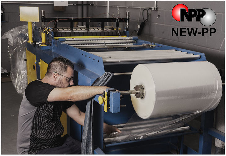 A imagem mostra um homem operando uma máquina industrial de grande porte, predominantemente nas cores azul e amarela. A máquina está processando um rolo grande de material plástico transparente, que está sendo desenrolado e alimentado através de diversos mecanismos. O homem, que usa óculos e uma camiseta cinza e preta, parece estar ajustando ou monitorando o processo. A máquina possui várias partes complexas, incluindo rolos, guias e um sistema que parece selar ou cortar o material plástico. Há também uma tela amarela com informações ou controles na parte superior esquerda da máquina.